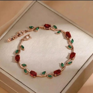 May include: A rose gold-toned bracelet featuring a vine-like chain design adorned with alternating oval-shaped red stones and marquise-cut green stones. The piece includes a metal clasp closure and is arranged in a circular shape on a flat surface.