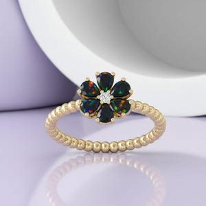 May include: A gold ring featuring a flower design with five black opal petals and a small diamond center. The ring band has a textured, beaded appearance. The ring is set against a soft purple background.