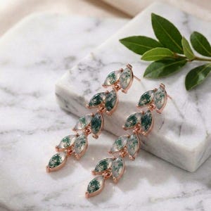 May include: Rose gold earrings with a leaf-like design. Each leaf shape is filled with a translucent green and white stone. The earrings are displayed on a marble surface. The design gives a natural and elegant look.