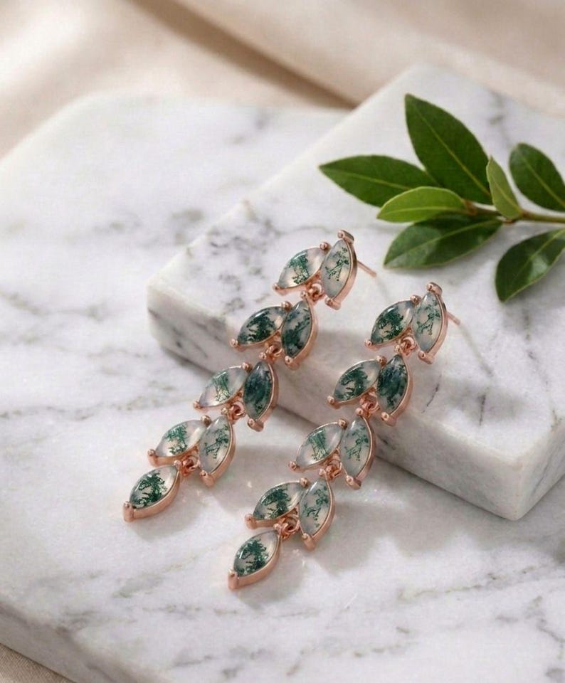 May include: Rose gold earrings with a leaf-like design. Each leaf shape is filled with a translucent green and white stone. The earrings are displayed on a marble surface. The design gives a natural and elegant look.