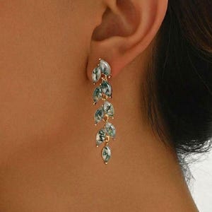 May include: Gold-coloured drop earrings with a leaf design. Each earring has multiple leaf-shaped pendants with a marbled green and white pattern. The earrings are shown on a person's ear.