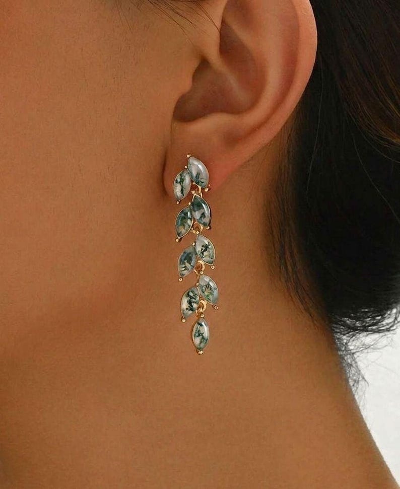 May include: Gold-coloured drop earrings with a leaf design. Each earring has multiple leaf-shaped pendants with a marbled green and white pattern. The earrings are shown on a person's ear.