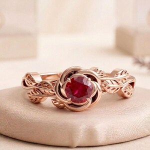 May include: A rose gold ring featuring a central round-cut deep red gemstone. The band is designed with intricate, sculpted leaf motifs that wrap around the finger, creating a botanical aesthetic that frames the vibrant red stone at the center.