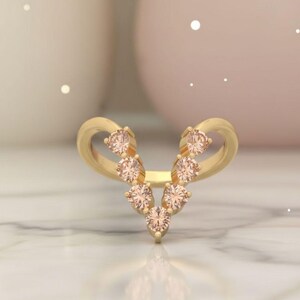 May include: A gold-colored ring with a V-shaped design. The ring features seven peach-colored gemstones. The ring's band curves upwards on both sides, creating a unique and elegant look. The ring is set against a soft, blurred background.