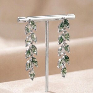 May include: Silver drop earrings with a leaf design. Each earring has multiple leaf-shaped pendants with a translucent look and green veining. The earrings are displayed on a silver stand. The earrings are a delicate piece of jewellery.