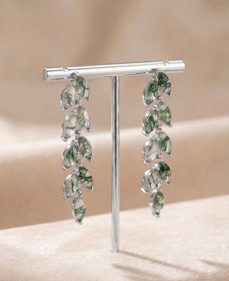 May include: Silver drop earrings with a leaf design. Each earring has multiple leaf-shaped pendants with a translucent look and green veining. The earrings are displayed on a silver stand. The earrings are a delicate piece of jewellery.