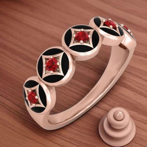 Peut inclure: Une bague en or rose ornée de cinq éléments circulaires noirs et dorés, chacun avec une pierre rouge centrale. La bague a un anneau lisse et un élément décoratif assorti.
