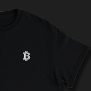 Op de afbeelding: Zwart T-shirt met ronde hals en een zilveren geborduurd Bitcoin-symbool op de borst. Het T-shirt heeft korte mouwen en een klassieke pasvorm. Een eenvoudig ontwerp voor cryptocurrency-liefhebbers.