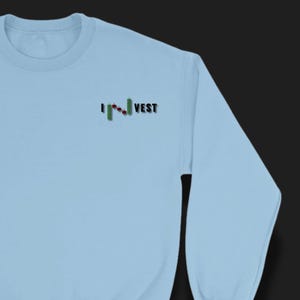 May include: Light blue sweatshirt with the word "I" and "VEST" in black text. A green and red stock market graph is between the words "I" and "VEST". The sweatshirt has a crew neck and long sleeves.