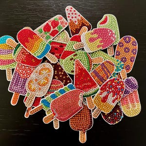 May include: A collection of assorted popsicle-shaped stickers decorated with colorful, sparkling rhinestones. Each sticker features a unique design, including patterns of fruit, swirls, and solid colors, all crafted with a faceted, gem-like texture.