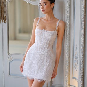 May include: A white mini dress featuring a corset-style bodice with intricate lace overlay, thin lace-trimmed shoulder straps, a square neckline, and a scalloped fringe hemline. The garment is constructed with structured vertical seams.
