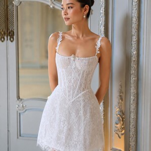 May include: A white lace mini dress featuring a corset-style bodice, thin floral-detailed straps, and a straight neckline with a small central embellishment. The garment has a fitted waist and a short, flared skirt with a delicate fringed hem.