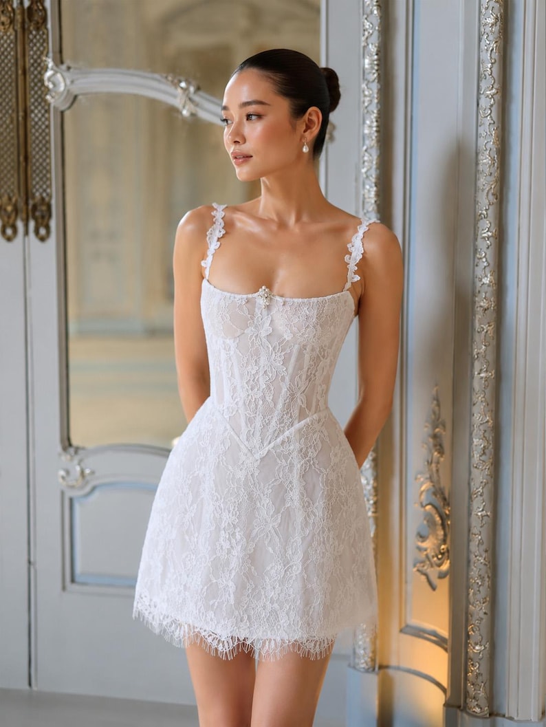 May include: A white lace mini dress featuring a corset-style bodice, thin floral-detailed straps, and a straight neckline with a small central embellishment. The garment has a fitted waist and a short, flared skirt with a delicate fringed hem.