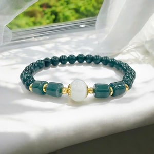 以下が含まれることがあります： 白い中心のビーズと金色のアクセントが付いた、濃い緑色のビーズブレスレット。 ブレスレットは、丸いビーズと長方形のビーズが混ざっています。 ブレスレットは白い表面に表示されています。