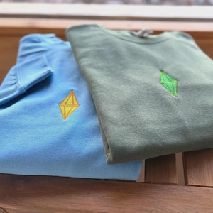 May include: Two folded sweatshirts, one light blue and one sage green. Each has a diamond-shaped embroidered design in yellow and green, respectively. The sweatshirts are made of a soft material, suitable for casual wear.
