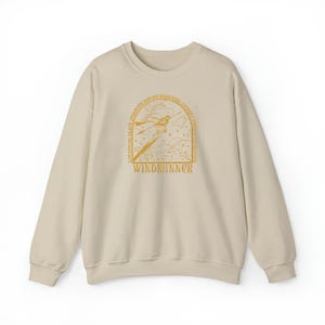 May include: A beige sweatshirt with a crew neck. The front features a gold illustration of a figure with a sword, surrounded by text that reads "WINDRUNNER" and other phrases. The design is centered on the chest.