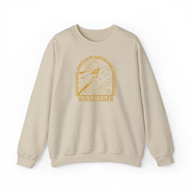 May include: A beige sweatshirt with a crew neck. The front features a gold illustration of a figure with a sword, surrounded by text that reads "WINDRUNNER" and other phrases. The design is centered on the chest.