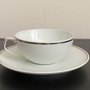 May include: White teacup and saucer set with a silver trim. The teacup has a curved handle and sits on a matching saucer. The set is made of porcelain and is suitable for serving tea or coffee.