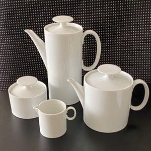 May include: A white porcelain tea set including a tall coffee pot, a teapot, a sugar bowl, and a creamer. The set has a minimalist design with rounded shapes and a glossy finish. The background is a black and white polka dot pattern.
