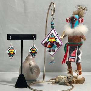 May include: A pair of beaded earrings and a beaded pendant, all featuring a geometric design with a central figure in a white, purple, orange, and blue color scheme. The earrings and pendant are hanging from a gold stand, and a small lizard figurine is in the foreground.