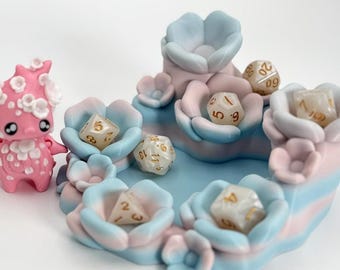 Puede incluir: Un juego de bandeja de dados y figura en colores pastel. La bandeja tiene forma de flores azules y rosas, con varios dados. Una figura rosa con detalles florales está al lado. Un regalo único para los entusiastas de los juegos de mesa.