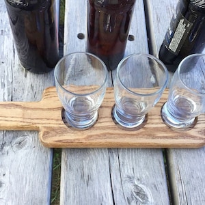 May include: A wooden beer flight tray with three empty glasses and a handle. The tray is made of light brown wood with dark brown streaks.