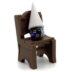 May include: A miniature brown wooden chair holds a blue and black dice with white numbers, topped with a white cone-shaped hat. The chair has a rustic design, and the dice is positioned as if it were a gnome.