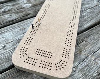 May include: A wooden game board with a circular track and holes for pegs. The board is brown and has a natural wood finish. There are three pegs on the board, two black and one gold.