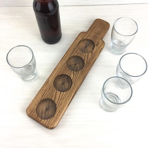 May include: Wooden beer flight tray with five slots for glasses. The tray is stained a dark brown color and has a natural wood grain. A brown glass beer bottle is on the left side of the image. Four clear glass beer glasses are on the right side of the image.