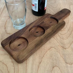 May include: A wooden serving tray with three circular indentations for holding glasses. The tray is made of dark brown wood and has a handle on one end.