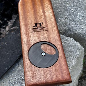 May include: A rectangular wooden bottle opener with a rounded top edge. The wood has a reddish-brown grain. A circular black textured insert is secured in the center with a silver screw. The text JT woodworks is printed in black on the wood.