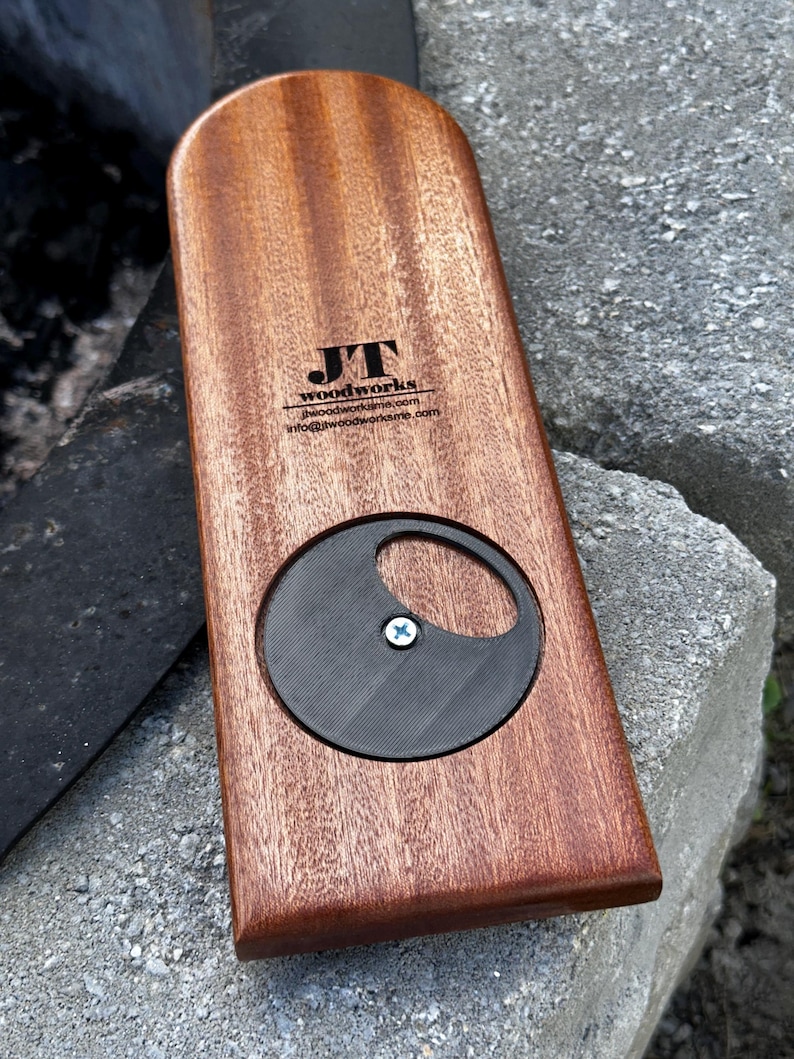 May include: A rectangular wooden bottle opener with a rounded top edge. The wood has a reddish-brown grain. A circular black textured insert is secured in the center with a silver screw. The text JT woodworks is printed in black on the wood.