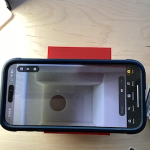 May include: A red phone stand with a smartphone on top. The phone is displaying a screen with a white background and a circular hole in the center. The screen also shows the words "Home, Search, Collection, Decks, and Scan".