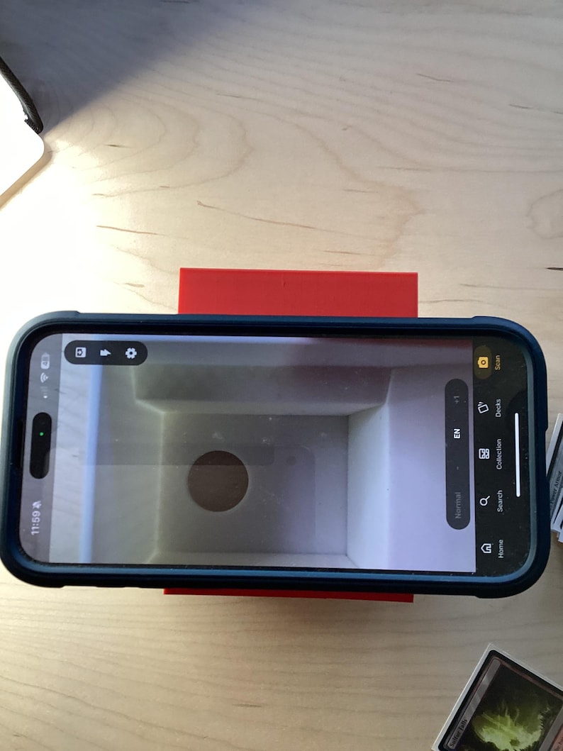 May include: A red phone stand with a smartphone on top. The phone is displaying a screen with a white background and a circular hole in the center. The screen also shows the words "Home, Search, Collection, Decks, and Scan".
