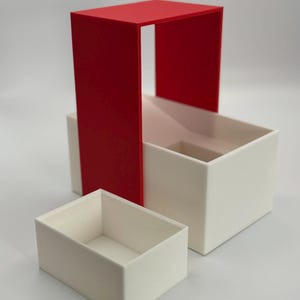 May include: A set of three white boxes with a red frame. The boxes are nested inside each other, with the largest box at the bottom and the smallest box at the top. The boxes are made of a smooth, white plastic.
