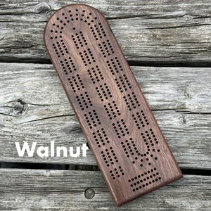 May include: A dark brown walnut wood cribbage board with a rounded top edge and a rectangular base. The board features a series of drilled holes arranged in a continuous track pattern for scoring. The word Walnut is printed in white on the board.