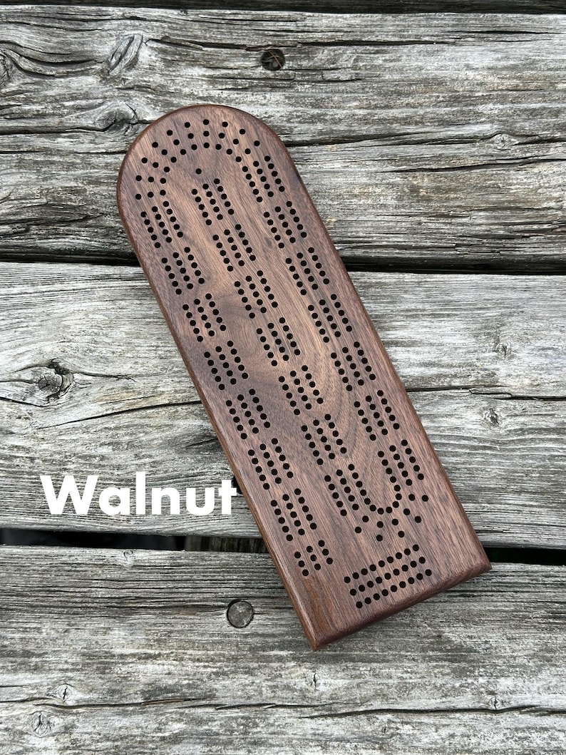 May include: A dark brown walnut wood cribbage board with a rounded top edge and a rectangular base. The board features a series of drilled holes arranged in a continuous track pattern for scoring. The word Walnut is printed in white on the board.