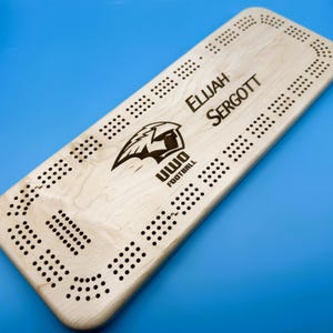 Peut inclure: Un jeu de cribbage en bois clair avec le nom "ELIJAH SERGOTT" et le logo "UWO FOOTBALL". Le jeu comporte de nombreux petits trous pour les chevilles et se détache sur un fond bleu.