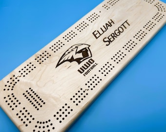 May include: A wooden cribbage board with a light finish, featuring the text "ELIJAH SERGOTT" and a logo for "UWO FOOTBALL". The board has numerous small holes for pegs and is set against a blue background.