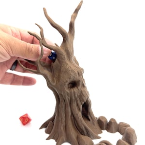 May include: A brown tree-shaped dice tower with a face carved into the trunk. A hand is placing a blue die into the tower. A red die is on the ground. The tower is designed for tabletop gaming.