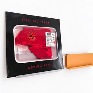 May include: A square Foo Fighters Medium Rare vinyl record cover featuring a graphic of a raw steak in a white plastic tray. The black cover has red text and a circular Foo Fighters logo on the meat. It rests on a silver metal wall shelf.