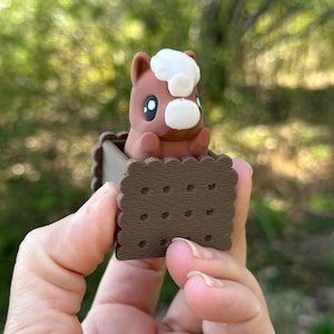 以下が含まれることがあります： 茶色と白の馬の置物が、茶色と白のクッキーの形をした箱の中に座っています。