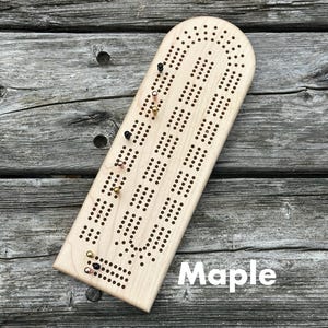 May include: A light-colored maple wood cribbage board with a rounded top and a series of drilled holes for scoring. The board features several metal pegs in black, copper, and gold colors inserted into the holes. The word Maple is printed.