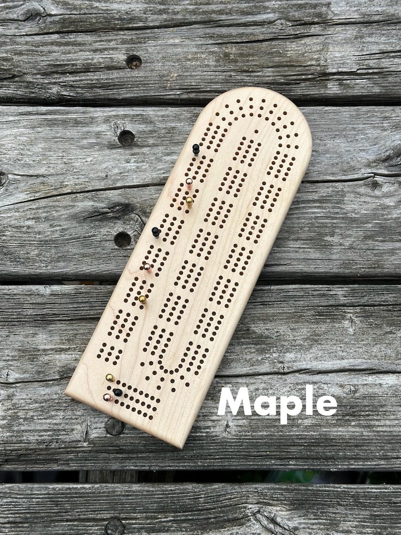 May include: A light-colored maple wood cribbage board with a rounded top and a series of drilled holes for scoring. The board features several metal pegs in black, copper, and gold colors inserted into the holes. The word Maple is printed.
