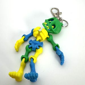 May include: A green, yellow and blue articulated skeleton keychain. The skeleton is posed with its arms outstretched and its legs bent. The keychain has a silver metal ring.