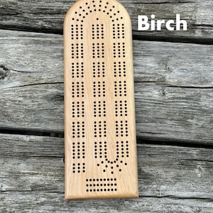 May include: A light-colored wooden cribbage board with a smooth finish and a rounded top edge. The board features a classic three-track layout with numerous drilled holes for scoring pegs. The word Birch is printed in white text on the right side.