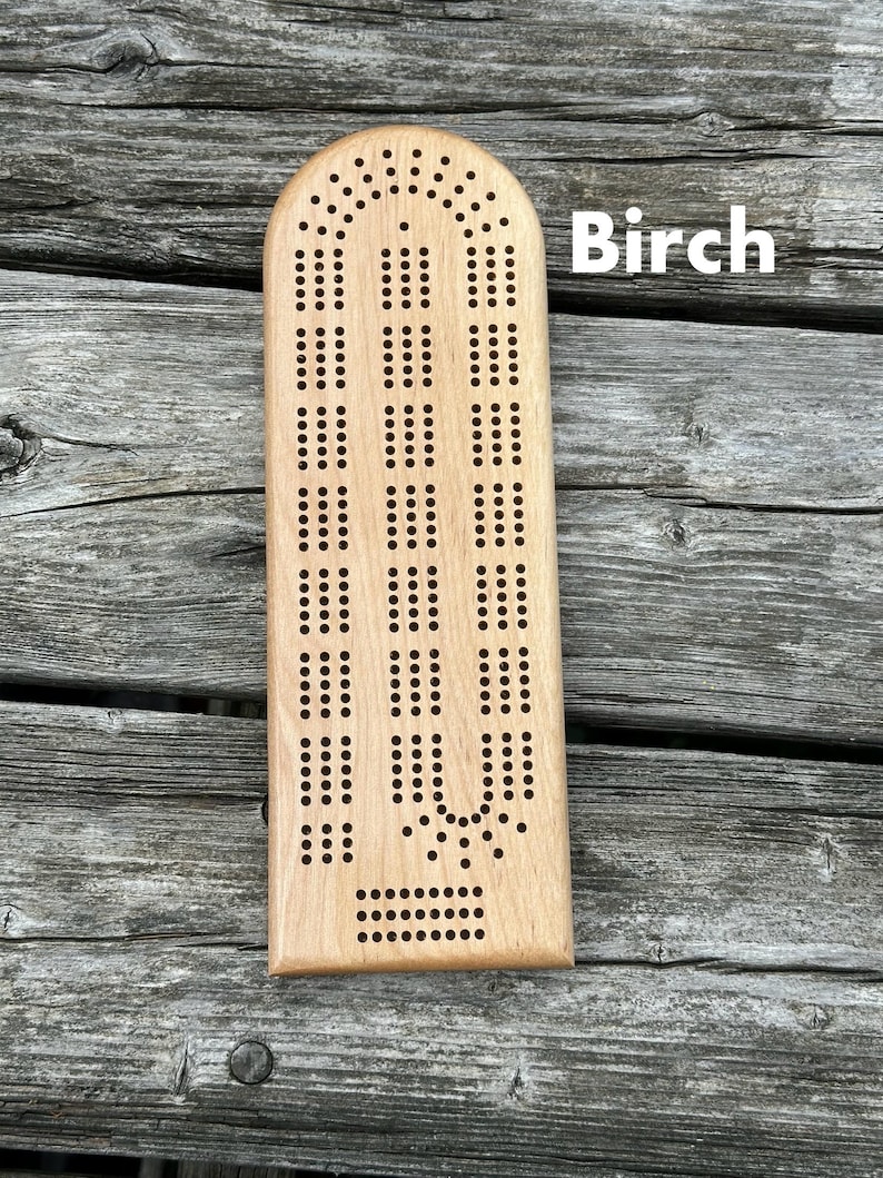 May include: A light-colored wooden cribbage board with a smooth finish and a rounded top edge. The board features a classic three-track layout with numerous drilled holes for scoring pegs. The word Birch is printed in white text on the right side.