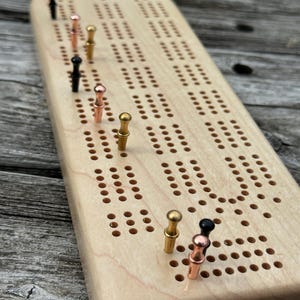 May include: A light-colored wooden cribbage board with a rounded end and multiple rows of drilled holes. The board features several metal pegs in black, copper, and gold tones inserted into various positions along the playing tracks.