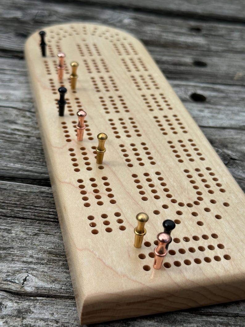 May include: A light-colored wooden cribbage board with a rounded end and multiple rows of drilled holes. The board features several metal pegs in black, copper, and gold tones inserted into various positions along the playing tracks.