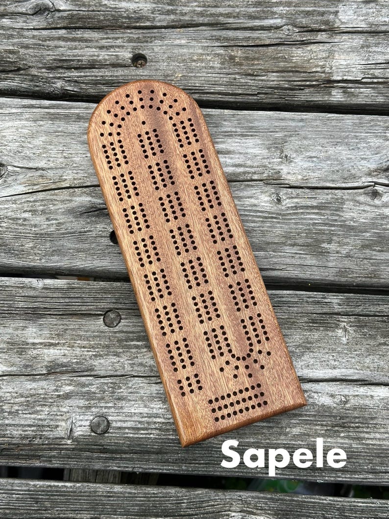 May include: A rectangular wooden cribbage board with a rounded top edge, featuring three parallel tracks of drilled holes for scoring. The board is crafted from brown Sapele wood with a visible grain pattern and includes the text Sapele.